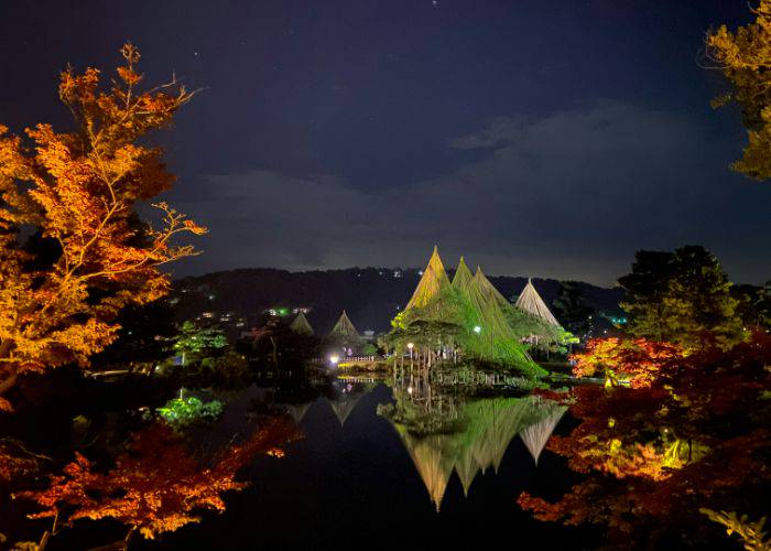 Kenrokuen Garden light up at night, with the orange leaves of trees in the foreground lit up.